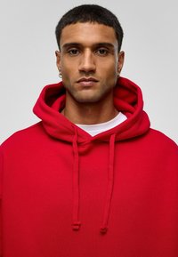 Rode hoodie met trekkoorden, zachte textuur, kangoeroezak, ribgebreide manchetten en zoom, gedragen over een wit shirt, model tegen een grijze achtergrond.