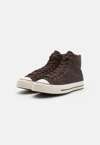 Hnědé vysoké tenisky vyrobené z texturovaného materiálu s bílými gumovými podrážkami a špičkami. Obsahují kontrastní černé pruhy podél podrážky.