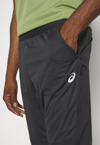 Sorte atletiske shorts med en glat tekstur, der har en lynlås-lomme og Asics-logoet i hvidt på venstre side.