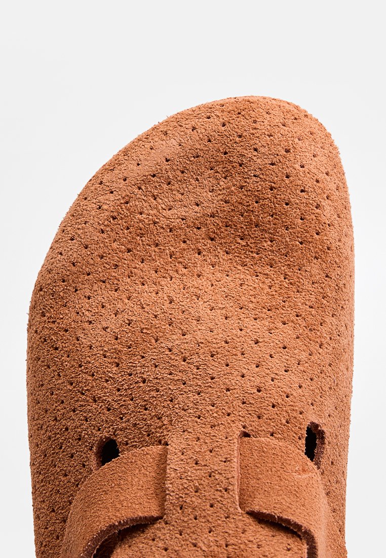 Nahaufnahme einer braunen Wildlederschuhspitze mit durchbrochenen Details und einem Riemen über der Vorderseite vor einem schlichten Hintergrund.