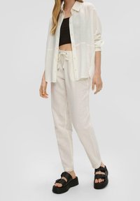 Lätt genomskinlig vit skjorta med knappar över en svart crop-top, i kombination med krämfärgade byxor med dragsko och svarta plattformssandaler.