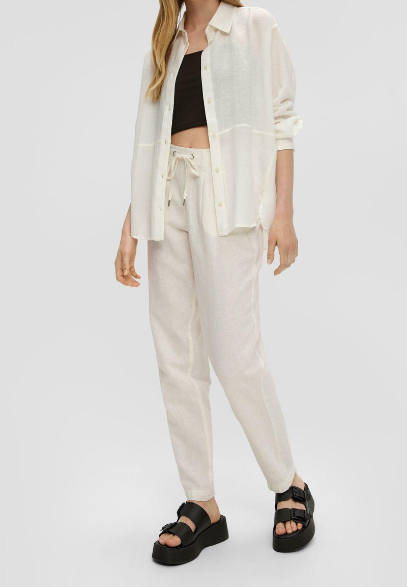 Lätt genomskinlig vit skjorta med knappar över en svart crop-top, i kombination med krämfärgade byxor med dragsko och svarta plattformssandaler.