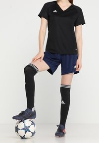 Jersey deportivo negro con cuello en V, pantalones cortos azules, calcetas negras hasta la rodilla con rayas blancas, y botas de fútbol sobre un balón de fútbol negro y blanco con un patrón.