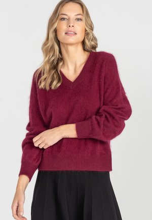 Kobieta z falującymi blond włosami, ubrana w miękki bordowy sweter w serek oraz czarną plisowaną spódnicę, na tle jasnego jednolitego tła.