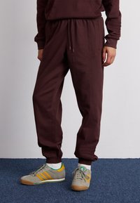 Burgundské tepláky s elastickým pasem, bočními kapsami a žebrovanými manžetami, doplněné šedými teniskami s zelenými a žlutými akcenty.