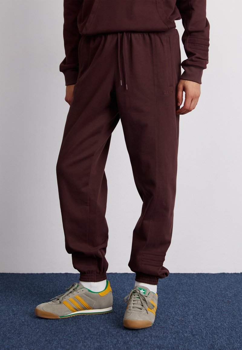 Burgundské tepláky s elastickým pasem, bočními kapsami a žebrovanými manžetami, doplněné šedými teniskami s zelenými a žlutými akcenty.