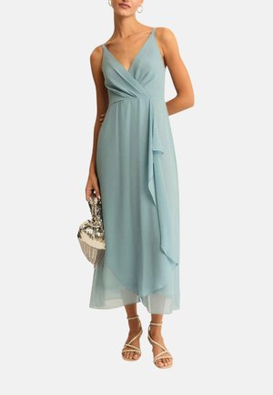 Donna che indossa un vestito midi azzurro chiaro senza maniche con scollo a V e dettaglio con volant, tiene una pochette argentata con paillettes e indossa sandali con cinturini.