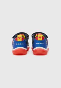 Baskets bleues et rouges avec une semelle en caoutchouc texturé, une sangle en velcro, des accents jaunes et un logo en relief. Présente un motif de super-héros au talon.