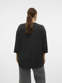 Haut noir ample avec des manches trois-quarts, en tissu texturé, à ourlet arrondi et avec un léger éclat, associé à un pantalon large gris.