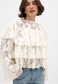 Lace blouse in cream with layered ruffles, sheer texture, and floral pattern. Features long sleeves and a round neckline. Paired with jeans.