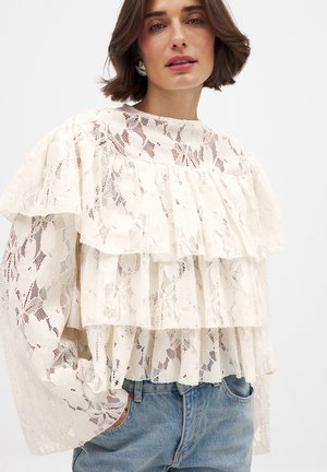 Lace blouse in cream with layered ruffles, sheer texture, and floral pattern. Features long sleeves and a round neckline. Paired with jeans.