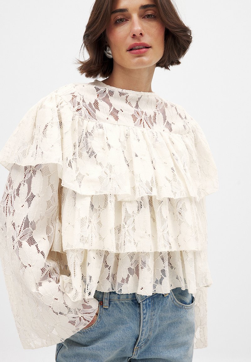 Lace blouse in cream with layered ruffles, sheer texture, and floral pattern. Features long sleeves and a round neckline. Paired with jeans.