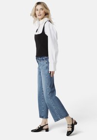 Haut sans manches noir sur une chemise blanche à col, associé à un jean droit bleu clair et des chaussures à talons slingback noirs en vernis avec des boucles.