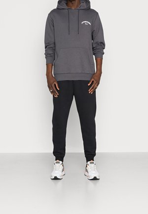 Haut à capuche gris avec poche avant et cordon de serrage, associé à un pantalon de survêtement noir et des baskets blanches avec des accents colorés.