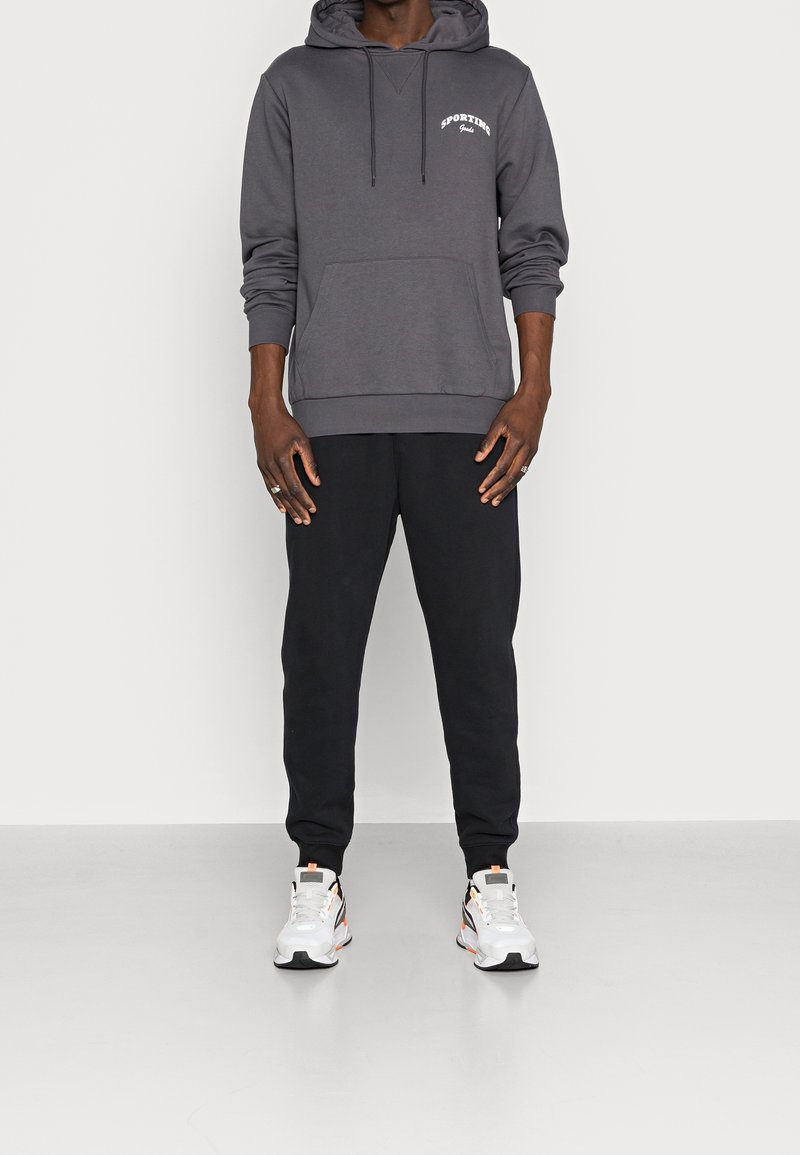 Felpa grigia con tasca frontale e cappuccio regolabile, abbinata a pantaloni sportivi neri e sneakers bianche con accenti colorati.