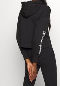 Sudadera corta negra hecha de tela suave, con una capucha grande y el logo blanco de "Champion" impreso en la manga izquierda.