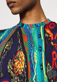 Närbild av en person som bär en flerfärgad stickad tröja med intrikata mönster i blå, röd, gul och grön ton.