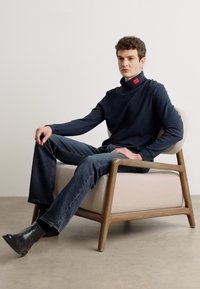 Maglione a collo alto navy in morbido tessuto, posato su una sedia moderna con struttura in legno e rivestimento beige chiaro, abbinato a jeans in denim scuro.
