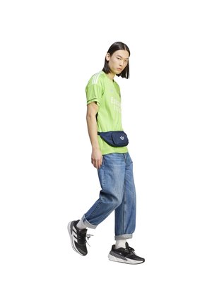 Grünes Sportshirt mit weißen Streifen, blauer Hüfttasche, lockeren blauen Jeans und schwarzen Sneakers mit weißen Akzenten.