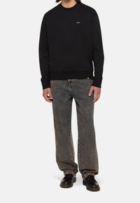 Personne portant un sweat-shirt noir, un jean large délavé à l'acide, et des bottes en cuir noires, debout devant un fond clair uni.