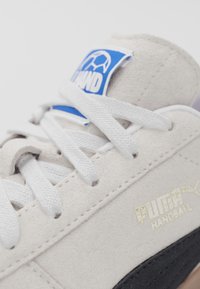 Scarpe da pallamano Puma con tomaia in suede grigio chiaro, accenti neri, lacci bianchi e un'etichetta logata blu con marchio dorato.