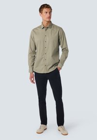 Olijfgroene button-up shirt met geometrisch patroon, lange mouwen, kraag en bruine knopen. In combinatie met zwarte broek en beige sneakers.