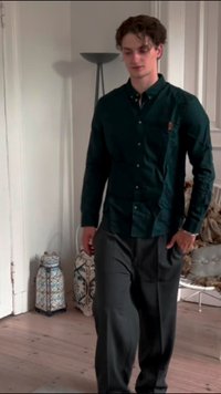 Camisa abotoada verde escura com um bolso, combinada com calças cinzentas largas. O ambiente apresenta lanterna decorativas e um chão de madeira clara.