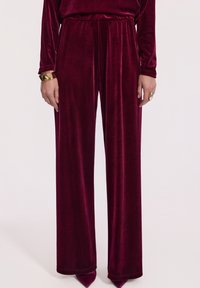 Kobieta ubrana w szerokie, burgundowe spodnie z aksamitnego materiału i dopasowaną bluzkę, dopełniona złotą bransoletką i dużym pierścionkiem.