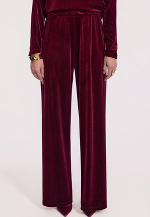 Kobieta ubrana w szerokie, burgundowe spodnie z aksamitnego materiału i dopasowaną bluzkę, dopełniona złotą bransoletką i dużym pierścionkiem.