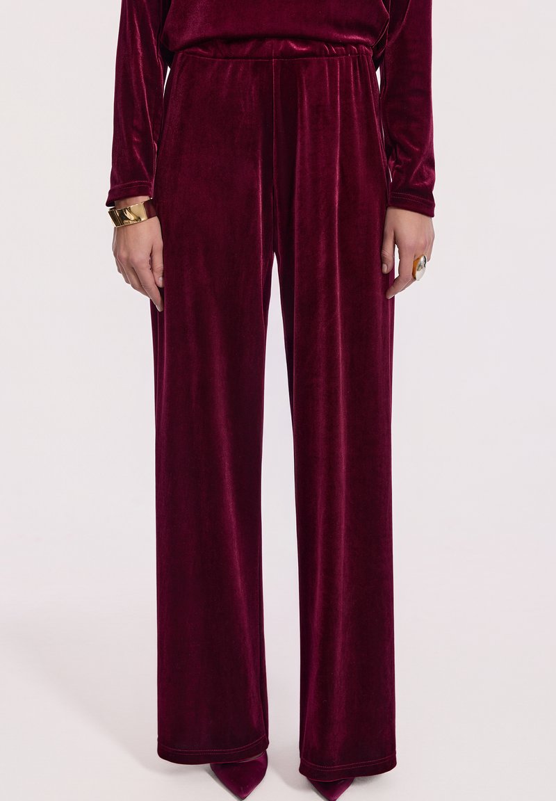 Kobieta ubrana w szerokie, burgundowe spodnie z aksamitnego materiału i dopasowaną bluzkę, dopełniona złotą bransoletką i dużym pierścionkiem.