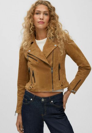 Kvinde med krøllet blondt hår iført en brun ruskindsjakke og mørkeblå jeans, stående mod en ensfarvet lys baggrund.