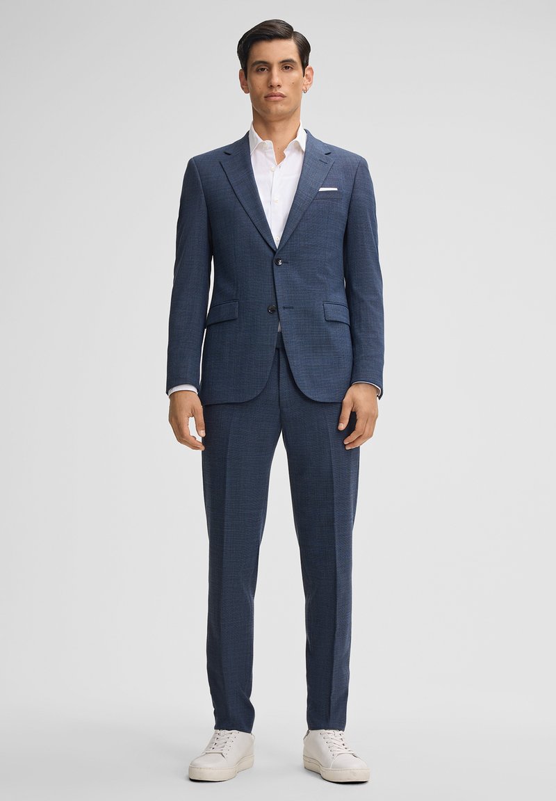 Completo blu navy in tessuto strutturato, con chiusura a due bottoni, rever a punta e due tasche frontali; abbinato a una camicia bianca e scarpe da ginnastica.