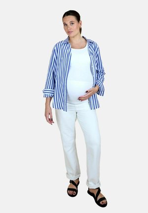 Gravid kvinde iført hvide bukser, hvid top, blåstribet skjorte og sorte sandaler, der blidt holder sin mave med den ene hånd.