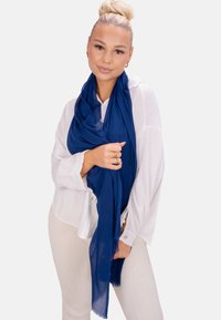 Écharpe bleu marine en tissu léger, drapée de manière décontractée. Portée avec un chemisier blanc texturé et un pantalon clair côtelé.