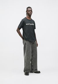 Μαύρο T-shirt με την επιγραφή «marimekko» σε λευκό, συνδυασμένο με φαρδιά, ανοιχτόγρια τζιν παντελόνια και μαύρα παπούτσια. Ουδέτερο φόντο.