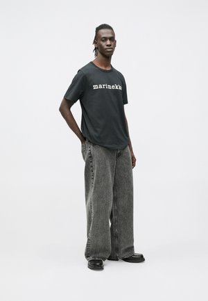 Juodas marškinėlis su baltu užrašu „marimekko“, derinamas su plačiomis, šviesiai pilkomis laisvo kirpimo kelnėmis ir juodais batais. Neutralus fonas.