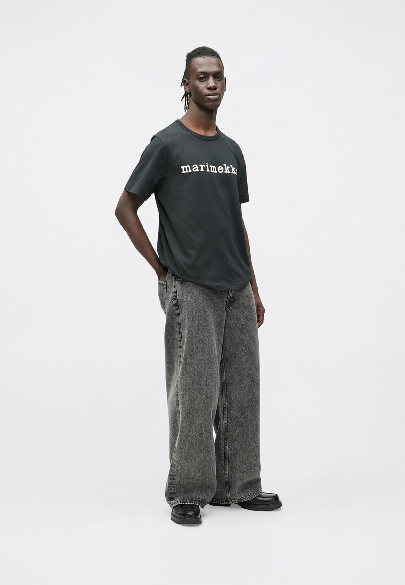 Μαύρο T-shirt με την επιγραφή «marimekko» σε λευκό, συνδυασμένο με φαρδιά, ανοιχτόγρια τζιν παντελόνια και μαύρα παπούτσια. Ουδέτερο φόντο.