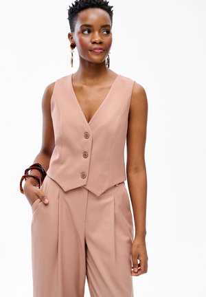 Kobieta w bezrękawniku w kolorze brzoskwiniowym zapinanym na guziki oraz pasujących szerokich spodniach, ozdobiona kolczykami-kołami i dużymi bransoletkami, patrząca na bok.