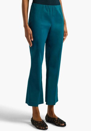 Donna che indossa pantaloni dritti color turchese con scarpe basse nere intrecciate, in piedi davanti a uno sfondo bianco uniforme.