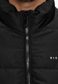 Chaqueta negra acolchada con cuello alto, con cierre de cremallera central, cremallera de tono plateado y la palabra "RICH" impresa en blanco.