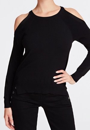 Kvinde iført sort skulderfri langærmet top og sorte bukser med den ene hånd på hoften foran en hvid baggrund.