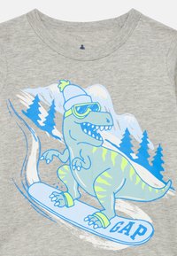 Γκρι βαμβακερό μπλουζάκι με ένα μπλε και πράσινο δεινόσαυρο που κάνει σνόουμπορντ, φοράει σκούφο και γυαλιά ηλίου, με γραφικά βουνών.