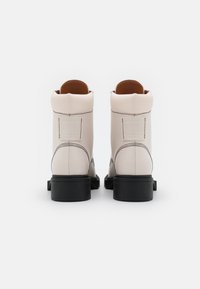 Bottes courtes en cuir blanc avec semelles en caoutchouc noires, présentant des coutures contrastées et un talon texturé. Comprend une étiquette de marque à l'arrière.