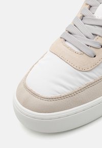 Sneaker featuring a beige suede and white padded upper, light gray laces, and a round, white rubber sole with textured detailing.