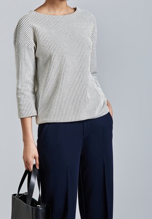 Frau trägt ein weiß gestreiftes Langarmoberteil und marineblaue Hose, hält eine schwarze Handtasche vor einem schlichten grauen Hintergrund.