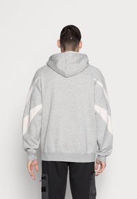 Ljusgrå hoodie med bakhuva, med vita snedställda paneler på ärmarna. Mjukt tyg med ribbade muddar och nederkant.