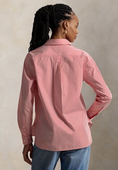 Γυναίκα με πλεξούδες, φοράει εφαρμοστό ροζ ριγέ πουκάμισο με μακριά μανίκια και μπλε τζιν, στέκεται με το ένα χέρι στη μέση, στραμμένη προς τα πίσω.