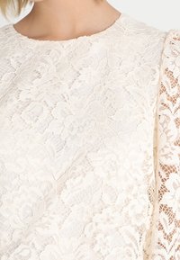 Top in pizzo color off-white con intricati motivi floreali e scollatura rotonda, dotato di maniche lunghe leggermente a sbuffo. Tessuto delicato e texturizzato.