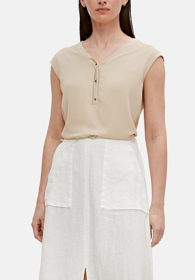 Femme portant un chemisier sans manches beige avec des boutons et une jupe blanche avec de grandes poches avant et une petite fente avant.