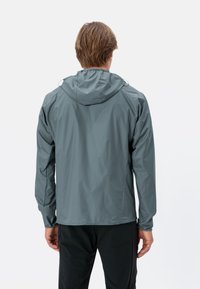 Giacca antipioggia leggera grigia con cappuccio, polsini elasticizzati e tessuto liscio e resistente all'acqua; presenta un design semplice e lineare.